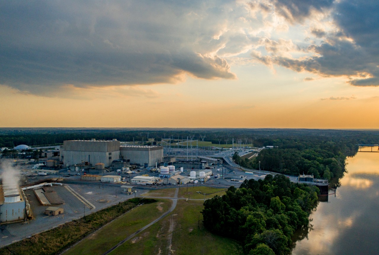 Southern Nuclear's Plant Hatch facility beside a calm river at sunset, with industrial buildings, power lines, and sky casting warm reflections.